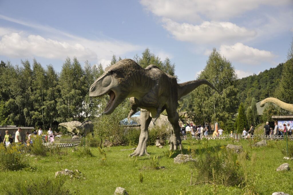 Model dużego dinozaura drapieżnego na łące w JuraParku w Bałtowie, w tle grupa zwiedzających i zalesione zbocze doliny